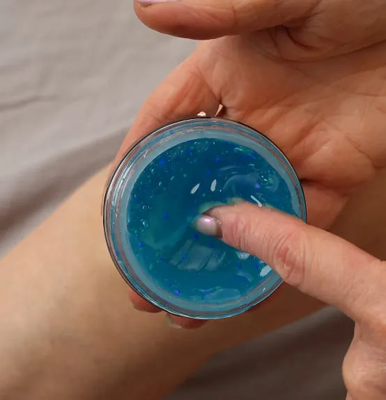 A finger taking Kinzeno massage gel from the open jar, showing the blue gel with suspended beads